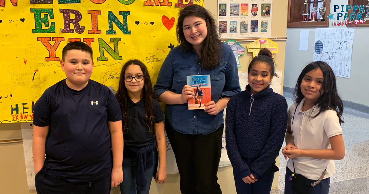 Erin Yun, the author of the Pippa Park series, is in the middle of the picture holding up her copy of Pippa Park Raises Her Game in a school hallway in front of a yellow poster board with her name on it. To each side of her are two middle-grade students smiling at the camera. 