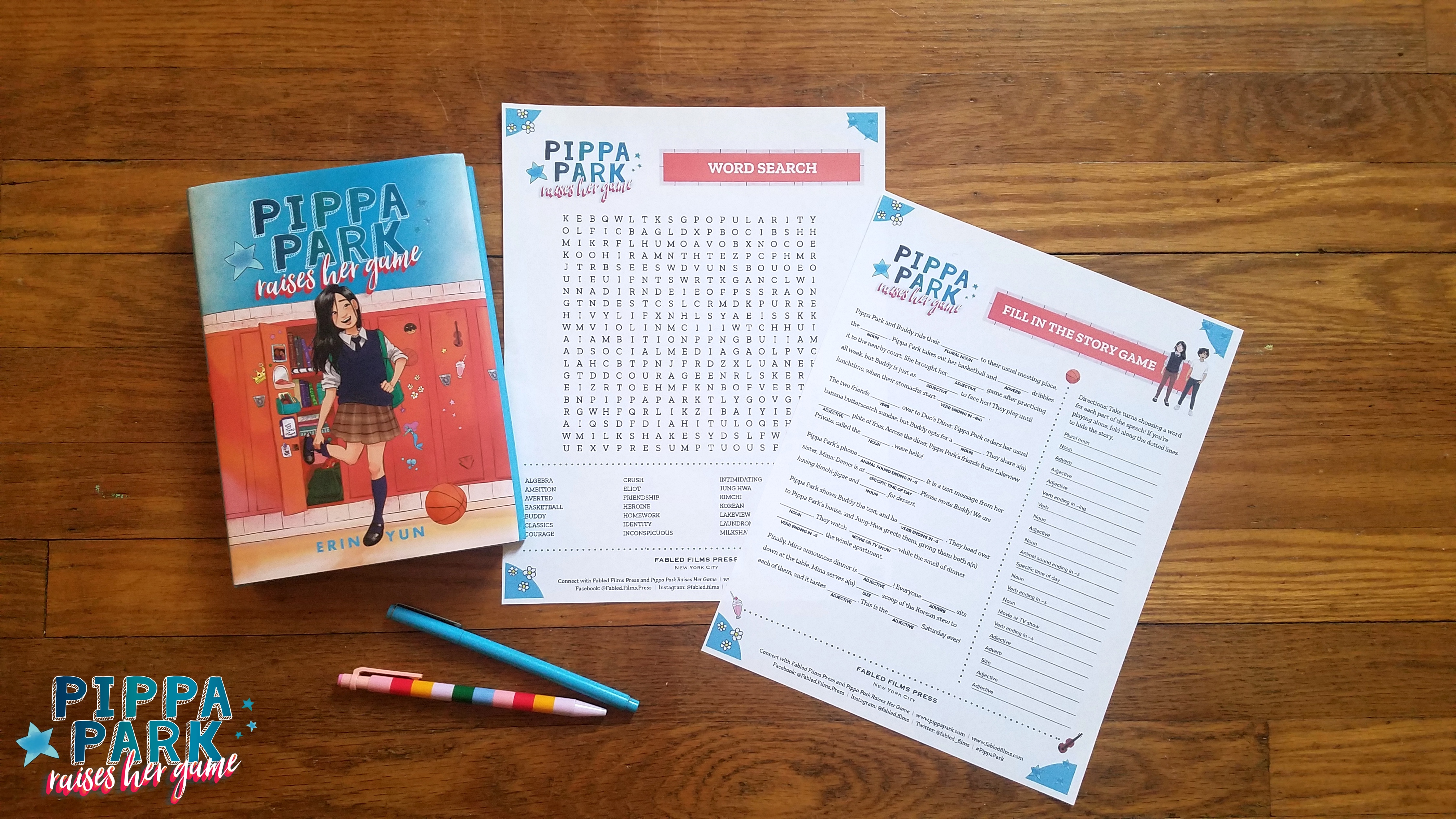 On a wooden desk is a copy of Pippa Park Raises Her Game and beside the copy are two Pippa Park writing activities: Word Search and Fill in the Story Game. Two pens are underneath the book copy and the writing activity printouts. 