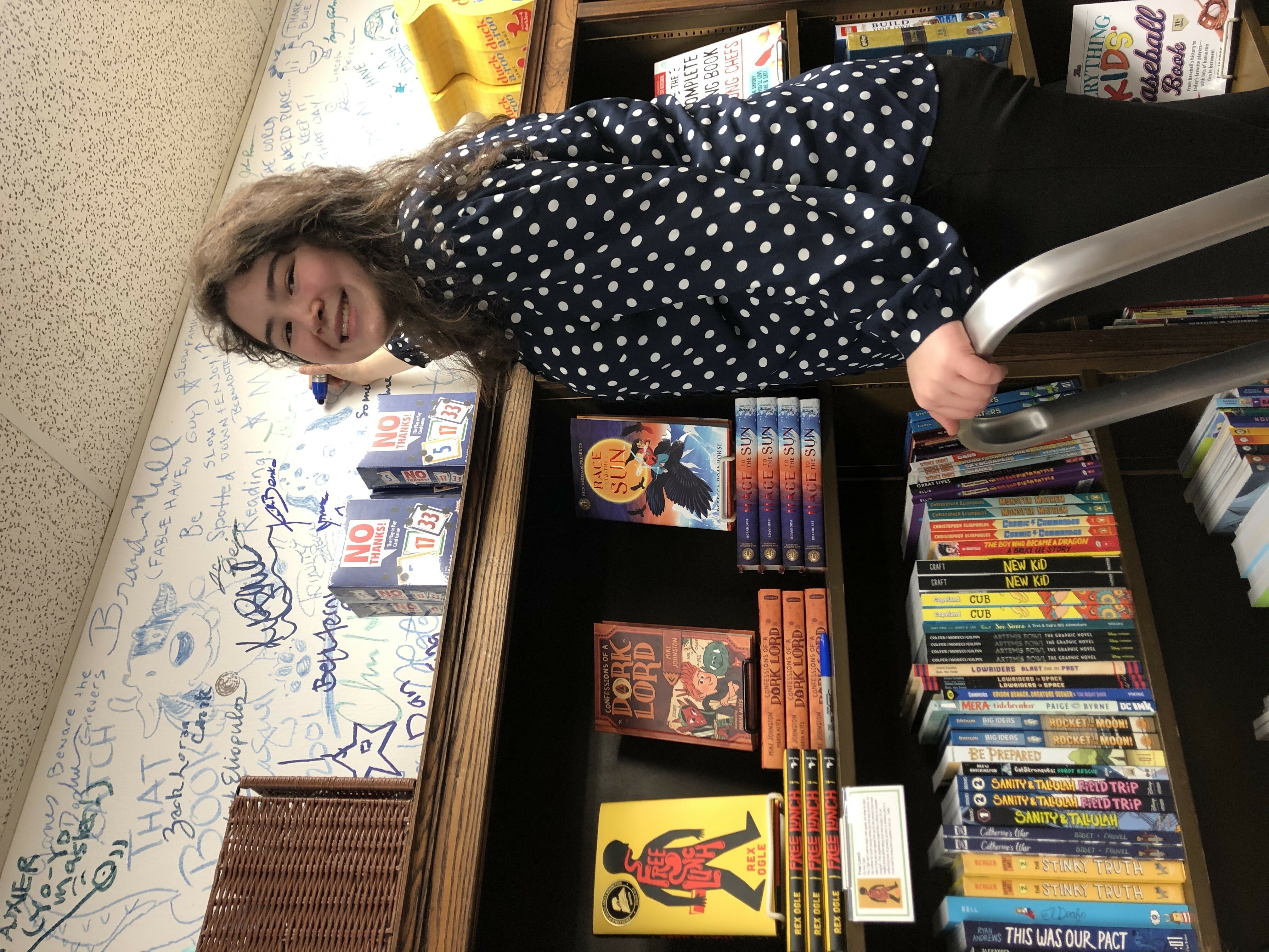 author Erin Yun smiles at the camera while signing a wall in a bookstore. The wall is covered in signatures.