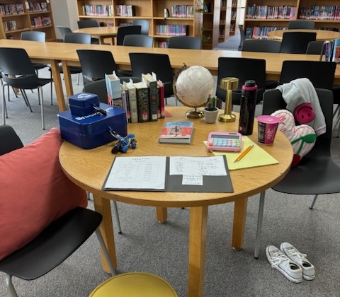 Table in library set up to play Pippa Park Escape Room with Breakout Edu Kit, homeworl folder, books, calculator, and other desk supplies.