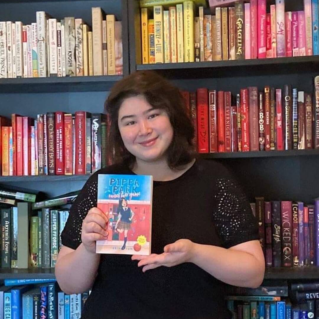 Erin Yun, the author of Pippa Park Raises Her Game, is holding up a copy of her book  in front of a rainbow organized bookshelf. 