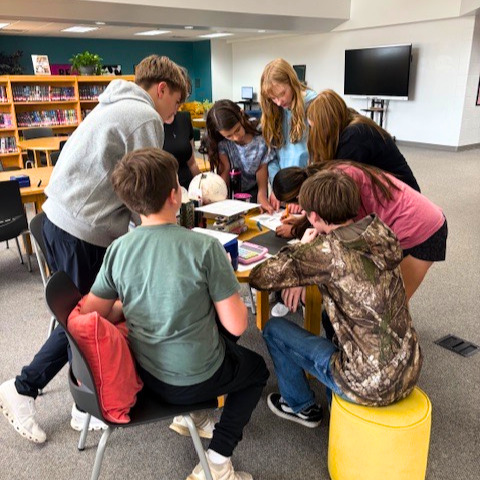 Middle School kids in a library surrounding a table where they are trying to solve the Pippa Park Escape Room Game
